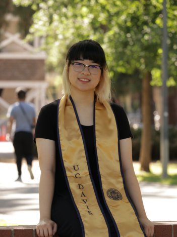 A young asian woman with dark brown hair with blonde ends wears a black dress and sits on a sign with a gold UC Davis stole around her neck and smiles at the camera.